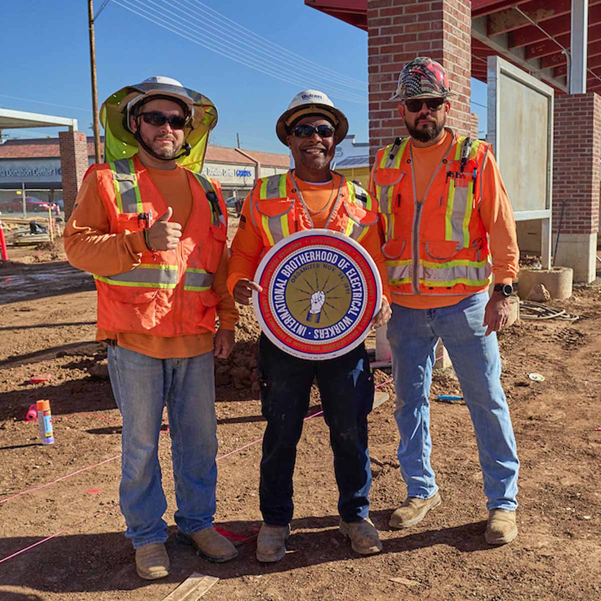 Three Electricians posing for the photo. They are on a jobsite. The middle person is holding up the IBEW logo and the first person is giving a thumb's up.